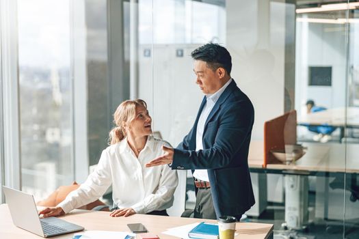 asian businessman is explaining plans to mature caucasian woman that working on laptop in office.