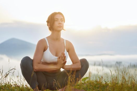 Front panoramic view of young female sitting in lotos pose in hills. Pretty woman doing yoga, looking forward with prayer hands. Concept of harmony with nature.