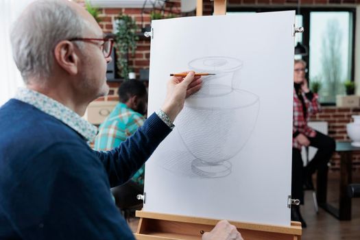 Elderly student drawing vase model on white canvas using sketching technique during art class in creativity studio. Diverse team attending creative lesson developing artistic skill