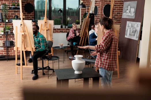 Painter teacher showing model for drawing course explaining illustration technique during art lesson in creativity studio. Multiethnic team sketching vase on painting canvas using graphic pencil