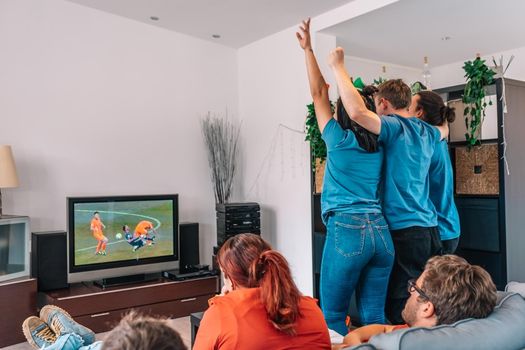 friends jumping for joy after their team's victory, watching football match on TV. opposing team sad after defeat leisure concept, three young adults in blue t-shirts. happy and cheerful. natural light in the living room at home.