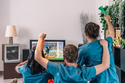 backs of friends cheering for their softball team while watching the game on TV on their living room couch. leisure concept, three young adults in blue t-shirts. happy and cheerful. natural light in the living room at home. trumpet