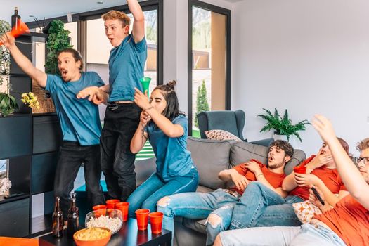 friends jumping for joy after their team's victory, watching football match on TV. opposing team sad after defeat leisure concept, three young adults in blue t-shirts. happy and cheerful. natural light in the living room at home.