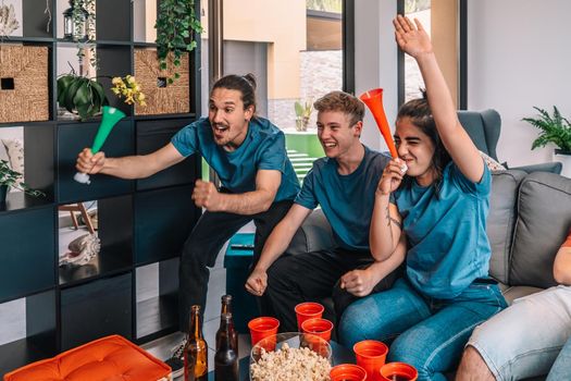 friends of championship fans shouting for joy after their team's victory, watching the broadcast on television. leisure concept, three young adults in blue t-shirts. happy and cheerful. natural light in the living room at home. trumpet