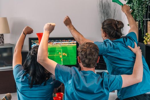 football friends celebrating a victory at home. young people watching sport on TV. leisure concept, three young adults in blue t-shirts. happy and cheerful. natural light in the living room at home.