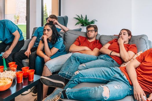 group of young people, bored watching a football match on television. tired youngster friends, at a house party. concept of leisure. concept of friendship. group of six people, friendly relationship, divided teams, red and blue, competitive.