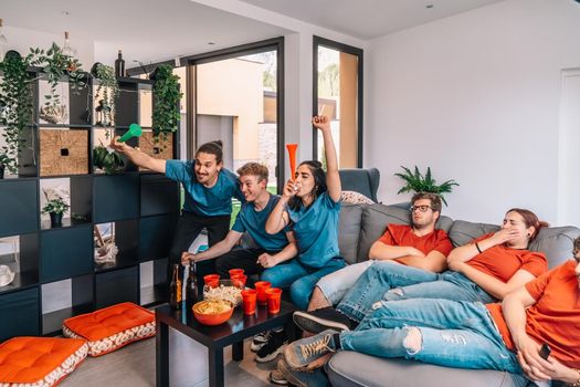 football fan group of friends celebrating a victory at home. young people watching sport on television. leisure concept. three young adults in blue jerseys and red jerseys. happy and cheerful. natural light in living room at home.
