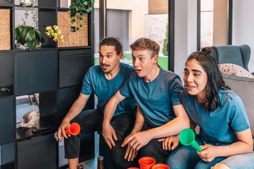 group of friends looking forward to the possible victory of their sports team, broadcast on television. leisure concept. three young adults in blue jerseys and red jerseys. happy and cheerful. natural light in living room at home.