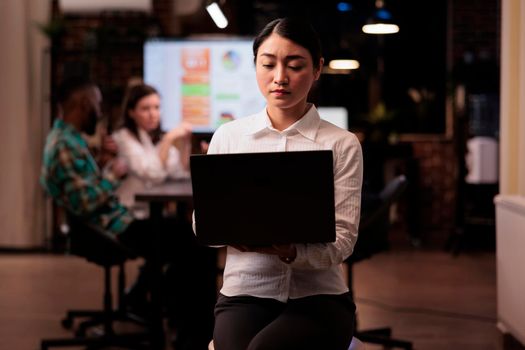 Portrait of asian woman holding laptop in startup office during late night meeting looking at screen. Employee posing confident working overtime with mixed team brainstorming marketing strategy.