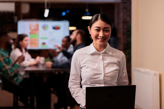 Portrait of smiling asian business employee holding laptop in startup office working overtime during late night meeting. Happy woman in white shirt posing confident with team doing group project.