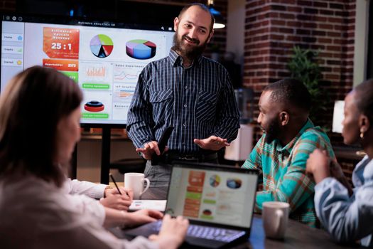 Startup employee happy with company sales results smiling at coworkers in late nigh meeting at office. Entrepreneur holding remote control presenting growing business revenue statistics on tv screen.