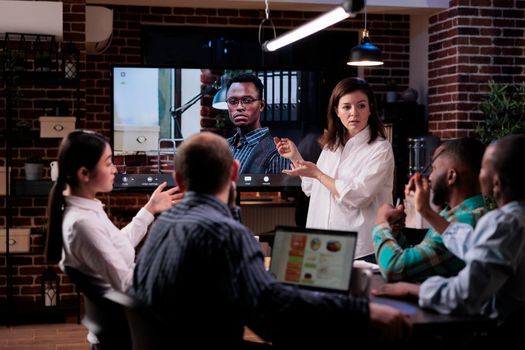 Caucasian business woman presenting in late night meeting with business owner in video call conference on big tv screen. Marketing team discussing strategy working overtime in startup office.