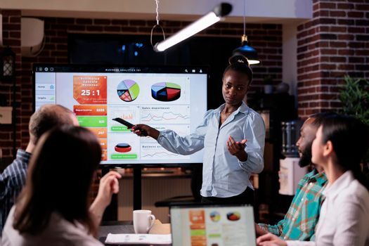 African american startup employee holding remote control presenting sales pie charts on tv screen in night meeting. Woman doing overtime at work talking about business strategy with diverse team.