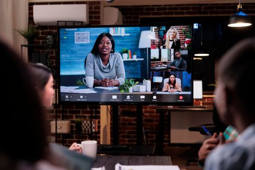 Selective focus on tv screen with african american business owner and clients in online video conference in startup office. Team in late night meeting working overtime brainstorming in internet call.