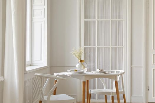 Glass vase with dried flowers and dishware placed on table near chairs and window in white spacious room at home