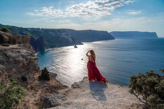 A girl with flowing hair in a long red dress stands on a rock above the sea. The stone can be seen in the sea. Sunny path to the sea from the sun