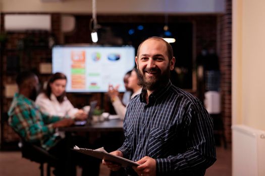 Portrait of smiling caucasian business employee holding clipboard in startup office working overtime during late night meeting. Happy man posing confident with team doing group marketing project.