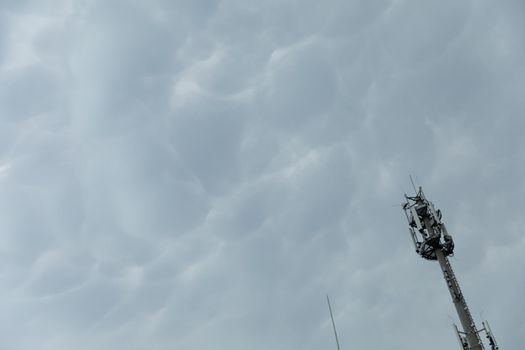 A tall pole with cell phone transmitters and receivers against a heavily overcast sky by an accumulation of bubble clouds.