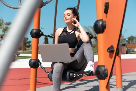the fitness girl in sportswear sitting on the playground in the summer outdoors with a laptop