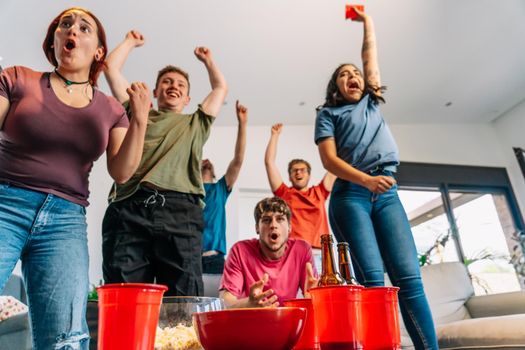 friends jumping for joy watching e-sports on TV after their team's victory. group of young people partying at home. leisure concept. happy and cheerful. natural light in the living room at home. trumpet