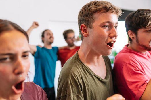 friends cheering for their team after scoring a point in their favour. sport broadcast on television. group of young people partying at home. leisure concept. three young adults in blue jerseys and red jerseys. happy and cheerful. natural light in living room at home.