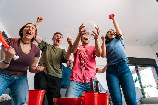 friends jumping for joy, throwing popcorn in the air after their team's victory. group of young people watching football at home. leisure concept. happy and cheerful. natural light in the living room at home. trumpet