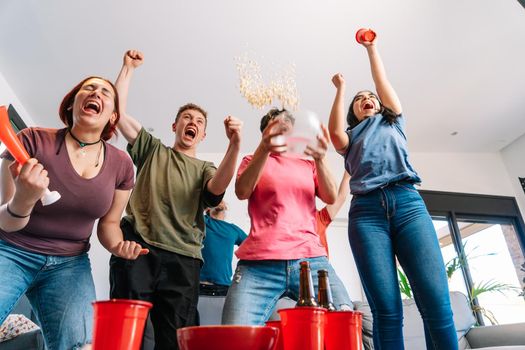 friends jumping for joy, throwing popcorn in the air after their team's victory. group of young people watching football at home. leisure concept. happy and cheerful. natural light in the living room at home. trumpet