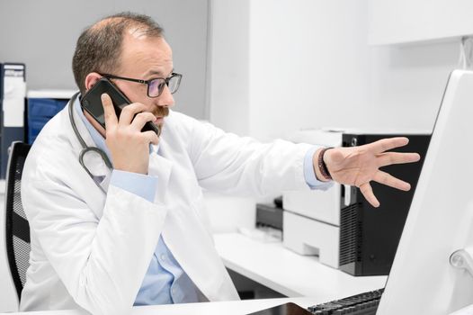 Male doctor pointing with finger on desktop computer while talking on the phone, discussing treatment with colleague. High quality photo