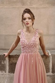 Beautiful brunette bridesmaid with luxury hairdo in gorgeous elegant stylish pale pink chiffon gown dress on flowers background. Wedding day in old beautiful European city.