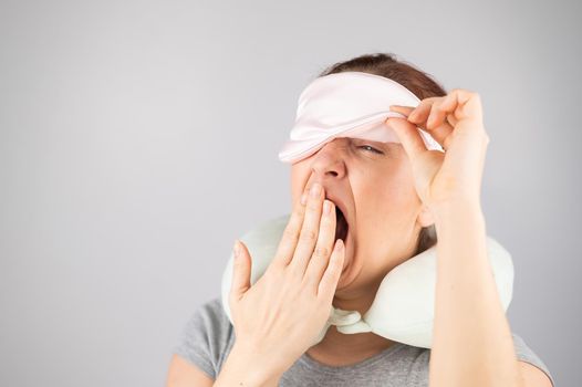 Caucasian woman yawns with travel pillow and sleep mask on white background