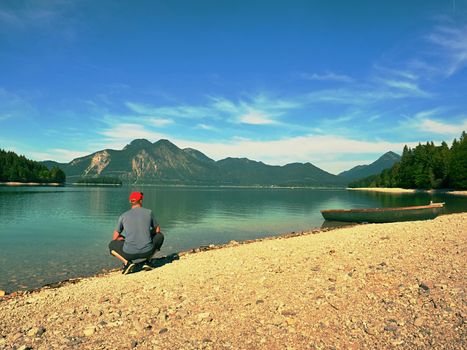 Man sitting on the stony bank of alpine mountain lake at anchored laminated boat. 