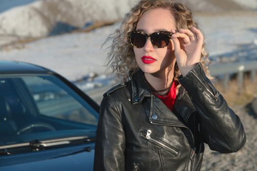 Street fashion portrait of young elegant luxury lady in sunglasses, curly hair and leather jacket posing near retro car. Copy, empty space for text. Holding on to sunglasses in the mountains.