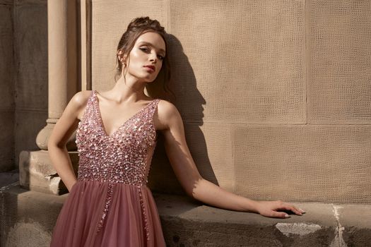 Beautiful brunette bridesmaid with luxury hairdo in gorgeous elegant stylish pale pink chiffon gown dress on flowers background. Wedding day in old beautiful European city.