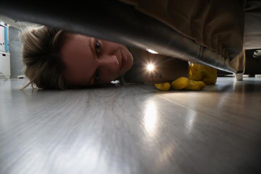 Close-up of woman using flashlight to look for something under bed. Female in protective gloves cleaning floor under bed