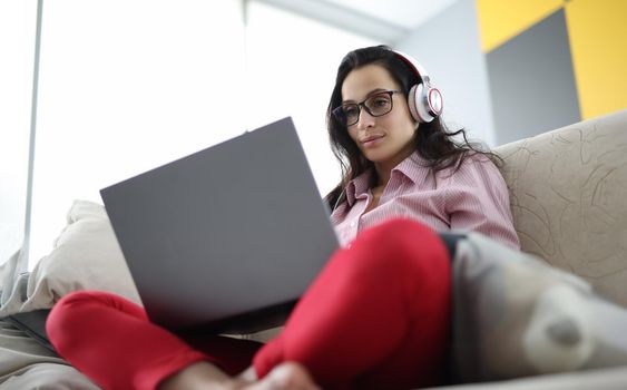 Portrait of woman sitting on sofa using laptop and headphones. Remote work or leisure time at home. Entertainment, distance learning or online meeting concept
