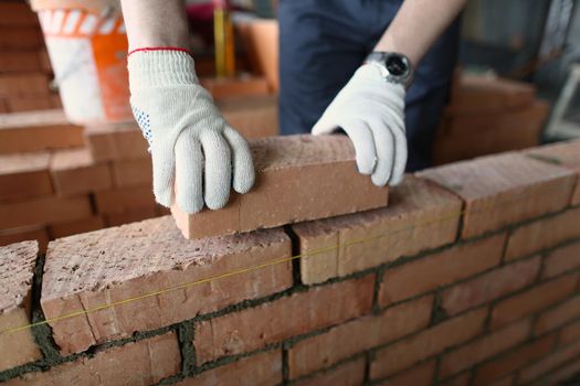 Close-up of male builder hands making make brickwork using red bricks. Builder building brick wall in house. Renovation and construction site concept