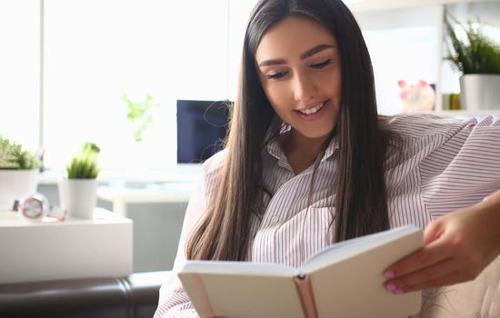 Portrait of young smiling woman reading book sitting on couch at home, interesting story. Cheerful brunette enjoy leisure time. Spare time and chilling concept