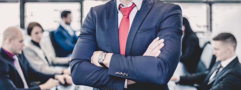 Successful team leader and business owner proudly standing with crossed arms with coworkers working in office in background. Business and entrepreneurship concept.