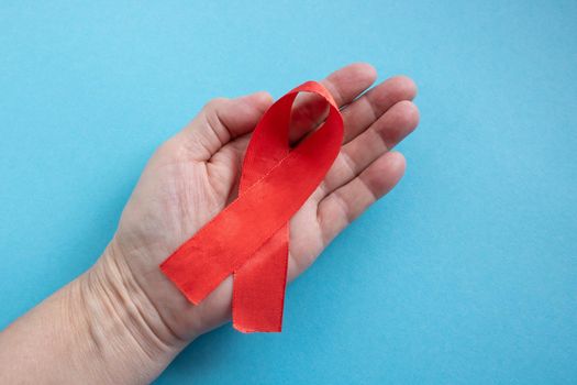 A hand holds a red ribbon on a blue background - the concept of World AIDS Day, Donor Day, Hemophilia Day.