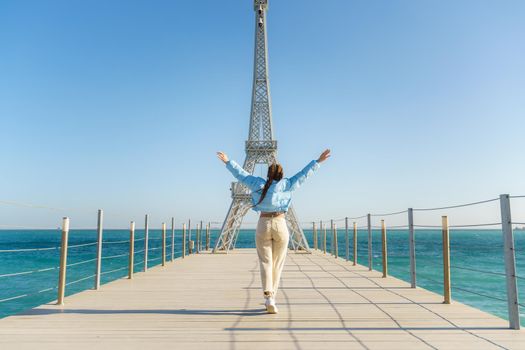 Large model of the Eiffel Tower on the beach. A woman walks along the pier towards the tower, wearing a blue jacket and white jeans