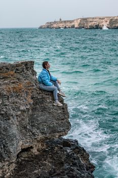 A woman in a blue jacket sits on a rock above a cliff above the sea, looking at the stormy ocean. Girl traveler rests, thinks, dreams, enjoys nature. Peace and calm landscape, windy weather