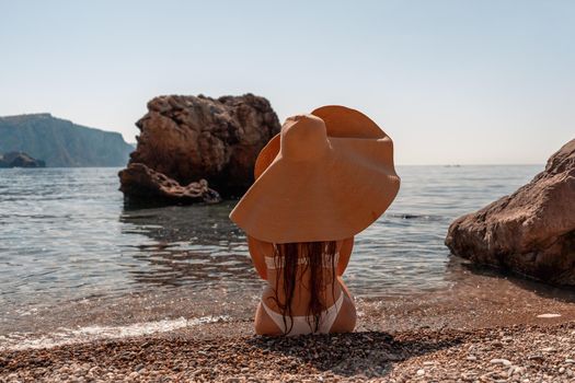 Tanned middle-aged woman with long hair and a white bathing suit. He sits on the seashore in a large sun hat with his back and looks at the sea