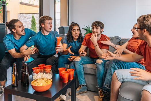 happy friends watching a sports event on TV, one of them unexpectedly changes the channel and makes the others angry. leisure concept. three young adults in blue jerseys and red jerseys. happy and cheerful. natural light in living room at home.