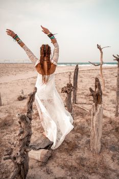 Model in boho style in a white long dress and silver jewelry on the beach. Her hair is braided, and there are many bracelets on her arms