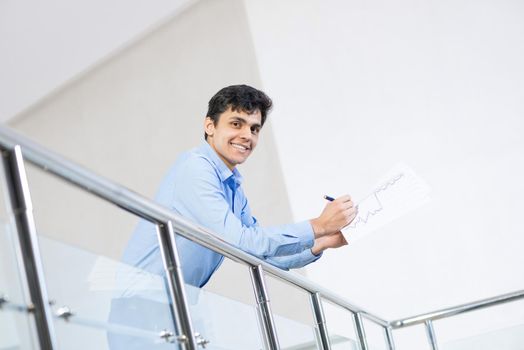 Young businessman. He holds documents in his hands, leans on the railing. He smiles and looks into the camera.