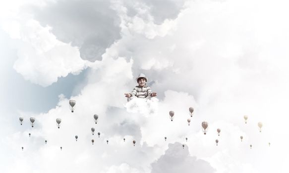 Young little boy keeping eyes closed and looking concentrated while meditating among flying balloons in the air with cloudy skyscape on background.
