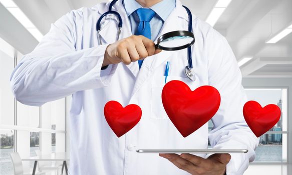 Conceptual image of professional doctor in white medical uniform is studying red heart symbol through magnifier while standing inside bright clinical building.