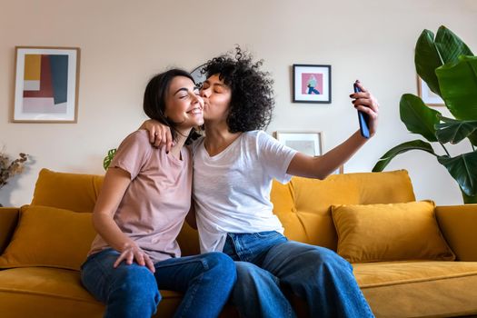 Young lesbian couple taking selfie at home using mobile phone. Girlfriend gives kiss on the cheek. LGTBQ and lifestyle concepts.