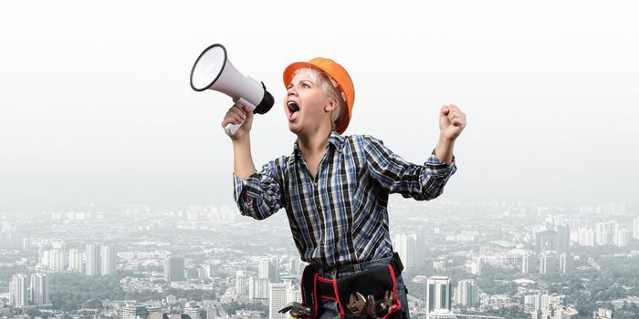 Expressive woman in safety helmet shouting into megaphone. Portrait of young emotional construction worker with wide open mouth on background of modern city. News announcement and advertisement.