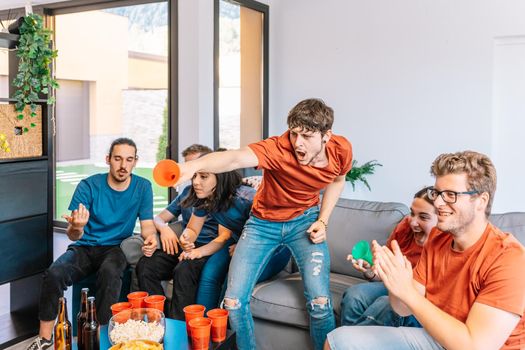 two groups of young friends facing each other watching a sports championship broadcast on TV. in the living room. leisure concept. happy and cheerful. natural light in the living room at home. trumpet